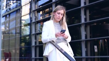 Kendine güvenen iş kadını cam ofis binasının yanındaki akıllı telefonu kullanarak mesajları ya da e-postaları, teknolojiyi, kurumsal yaşam tarzını ve profesyonel iş bağlantılarını kontrol ediyor.