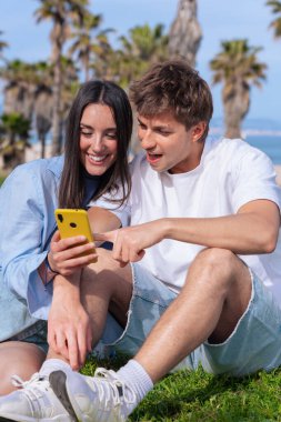 Palmiye ağaçlarının altında deniz kenarında oturan genç çift, birlikte sarı bir akıllı telefona bakarken gülümsüyor, ekrana işaret eden erkek arkadaş, aşkı, seyahat anlarını ve bugün dijital bağlanmayı yakalıyor. 