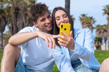 Palmiye ağaçlarının altında deniz kenarında oturan genç çift, birlikte sarı bir akıllı telefona bakarken gülümsüyor, ekrana işaret eden erkek arkadaş, aşkı, seyahat anlarını ve bugün dijital bağlanmayı yakalıyor. 