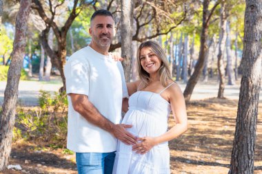 Güneşli ormanda birlikte kameraya bakan mutlu hamile çift, annelik kavramı, aile sevgisi, ebeveynlik, beklenti, gelecek hayat, hamilelik sevinci, bakım, birlik ve duygusal bağ..