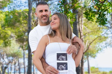 Mutlu hamile çift yeşil parkta ultrason fotoğrafı çekiyor, sarılıyor ve tutuyor. Annelik, sevgi, ebeveynlik, aile bağı, beklenti ve bebek beklerken doğa ve yeni hayatın tadını çıkarıyor.