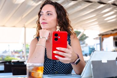 Çekici bir kadın kırmızı akıllı telefonu tutuyor ve açık hava masasında tablet ve içkiyle otururken yan bakıyor, modern yaşam tarzı, dijital iletişim, çevrimiçi çalışma, günlük iş tarzı