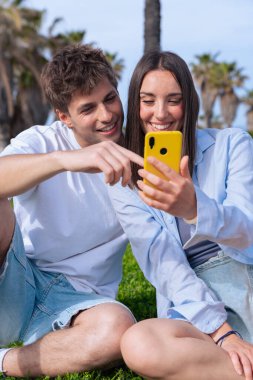 Palmiye ağaçlarının altında deniz kenarında oturan genç çift, birlikte sarı bir akıllı telefona bakarken gülümsüyor, ekrana işaret eden erkek arkadaş, aşkı, seyahat anlarını ve bugün dijital bağlanmayı yakalıyor. 