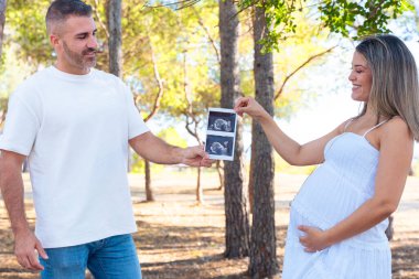 Güneşli parkta ultrason fotoğrafı tutan mutlu hamile çift annelik, ebeveynlik, aşk, beklenti, aile mutluluğu ve doğanın tadını çıkarırken bebek sahibi olmak..