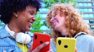 Güneşli bir park bankında sarı ve kırmızı telefonları karıştıran iki çok ırklı arkadaş viral videoları, sosyal medya güncellemelerini ve birlikte neşeli dijital yaşam tarzı anlarını paylaşıyorlar.. 