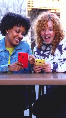 Mutlu çok ırklı genç kadınlar dışarıda birlikte oturuyorlar. Neşeli anları paylaşıyorlar. Sosyal medya bağlantıları için akıllı telefonları kullanıyorlar.