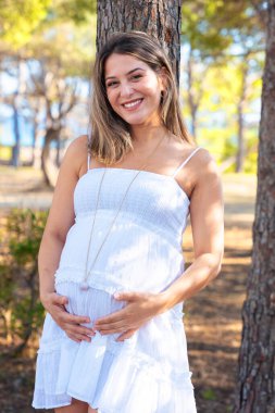 Ağacın yanında duran, gülümseyen ve karnına dokunan beyaz elbiseli mutlu hamile kadın. Hamilelik, doğa, şefkat, aşk, beklenti ve gün ışığında huzurlu bir an..
