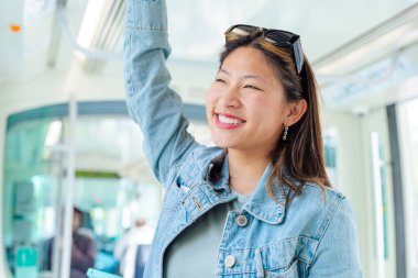 Toplu taşıma aracının içinde elinde tırabzan tutan gülümseyen kadın. Şehirde gidip gelen mutlu genç bayan, yolculuğunun tadını çıkarıyor, pozitif ruh hali ve günlük ulaşımı temsil ediyor..