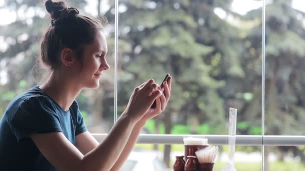 Használ smartphone az ablak mellett ülő nő — Stock videók