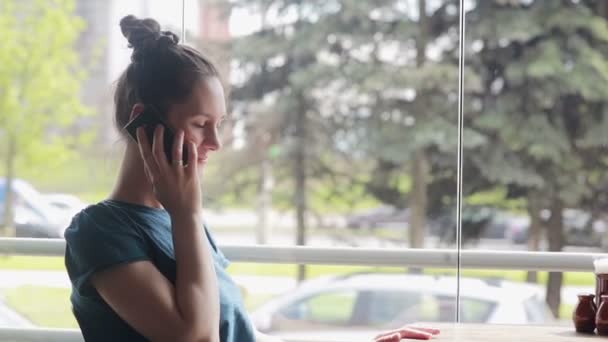 窓の近くに座って携帯電話で話している女性 — ストック動画