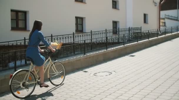 Молодая женщина в платье исследуя город на велосипеде с цветами в корзине в летнее время, медленная мама steadicam выстрел — стоковое видео