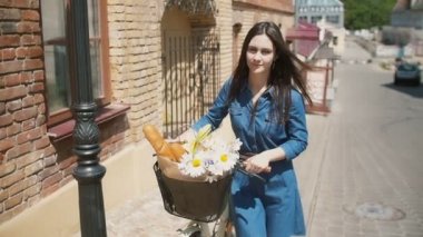 Kameraya gülümseyen esmer kız bir sepet içinde çiçek ve Fransız ekmeği ile bisiklet yürüyor, yavaş mo, steadicam atış