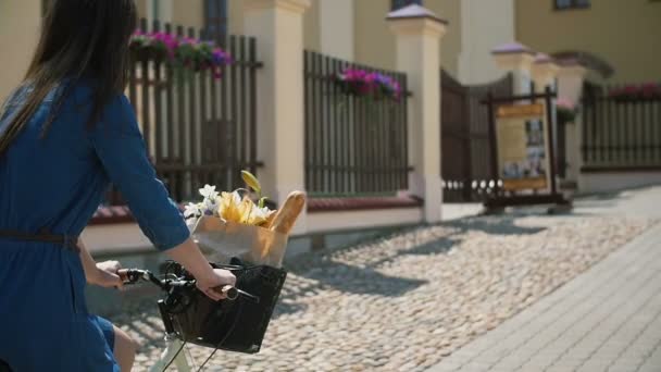 Fille en vélo dans la ville avec des fleurs et du pain français dans un sac en papier dans un panier, slow mo, steadicam shot — Video