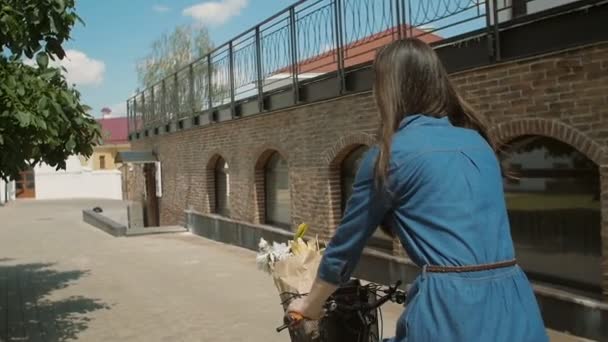 Ağaç yaprakları dokunmadan bir sepet içinde çiçek ile bisiklete binen bir elbise bir kızın geri görünümü, yavaş mo, steadicam atış — Stok video