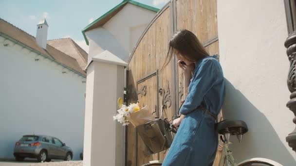 Joyeux brunette fille rire et parler au téléphone debout près de la vieille porte avec vélo et fleurs dans un panier, lent mo — Video