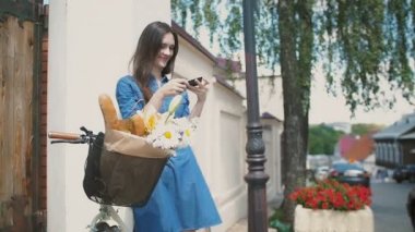 Esmer gülümseyen kız bir sepet içinde çiçekleri ile bir bisiklet ile binanın yanında duran onun akıllı telefon kullanarak, yavaş mo