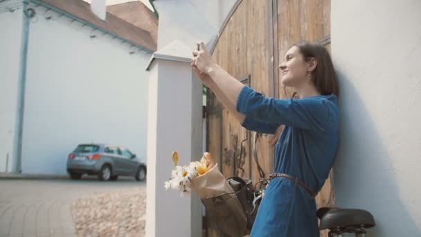 ブルネットの女の子は、自分撮りを取って、バスケットに花を持つ自転車で古い建物の近くに立って、遅いmo — ストック動画