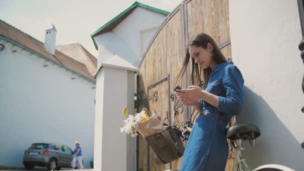 快乐的黑发女孩笑着，在电话上说话与自行车和鲜花在篮子里，使用她的智能手机，慢莫 — 图库视频影像