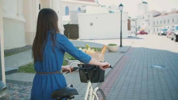 Vue arrière d'une fille aux cheveux ondulés marchant son vélo avec des fleurs et du pain dans un panier, slow mo, steadicam shot — Video