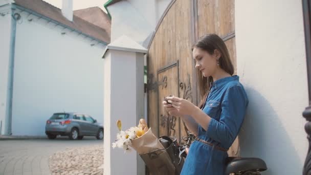 Brunetka dziewczyna za pomocą swojego smartfona stoi w pobliżu budynku z rowerem z kwiatami w koszyku, powolny mo — Wideo stockowe