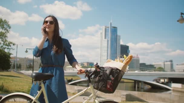 Belle fille debout dans la rue avec un vélo avec panier parlant au téléphone et souriant, 4k — Video