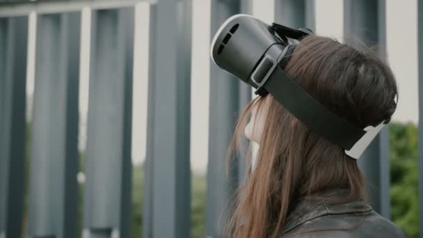 Menina morena com cabelo ondulado usa óculos de realidade virtual no espaço urbano. 4k — Vídeo de Stock