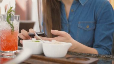Esmer kız akıllı telefon bir kafede dışarıda otururken bir saman tüpünden serin bir içki içme kullanır. 4k,