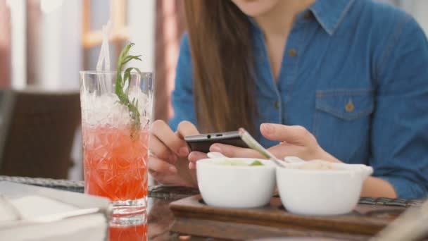 Chica morena utiliza el teléfono inteligente mientras está sentado fuera en un café caliente día de verano. 4k , — Vídeo de stock