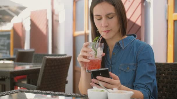 Menina morena usa telefone inteligente enquanto sentado fora em um café, beber e desfrutar de uma bebida fresca de tubo de palha. 4k , — Vídeo de Stock