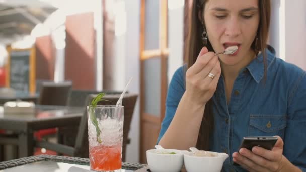 Brunette meisje gebruikt slimme telefoon zittend buiten in een café, het eten van ijs. 4k, — Stockvideo