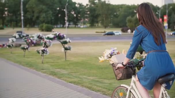 Vista lateral y trasera de una chica montando una bicicleta cerca de coches conducidos con flores en una cesta, lento mo, steadicam tiro — Vídeos de Stock