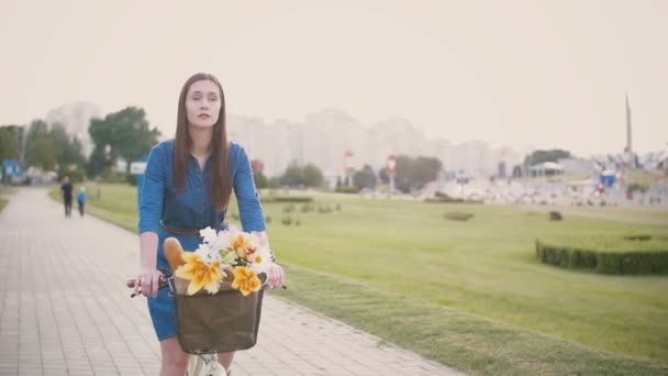 Morena menina em uma bicicleta com flores e pão francês em uma cesta enquanto pedalava na cidade, slow mo, steadicam tiro — Vídeo de Stock