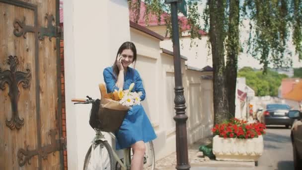 Девушка, стоящая на улице с велосипедом с корзиной, разговаривает по телефону и улыбается, 4k — стоковое видео