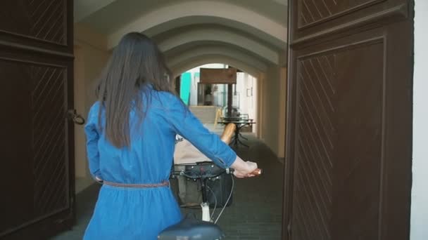 Vista trasera de una chica morena con el pelo largo caminando con una bicicleta en la puerta vieja en el casco antiguo, lento mo, steadicam shot — Vídeos de Stock