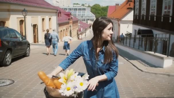 Chica camina su bicicleta por la calle con flores en la cesta como el sol brilla, coches aparcados cerca, lento mo, steadicam tiro — Vídeos de Stock