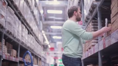 Bir adam karton kutulariçinde mal ile raflar yanında duruyor ve bir depoda listesini kontrol