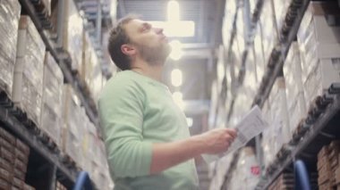 Bir adam mallarla rafların yanında duruyor, listesini kontrol ediyor, bir depoda gerekli şeyleri arıyor.