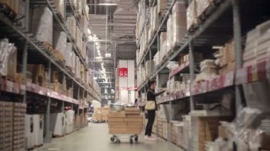 Young woman is looking for something on the shelves with goods in cardboard boxes between palettes in storage warehouse
