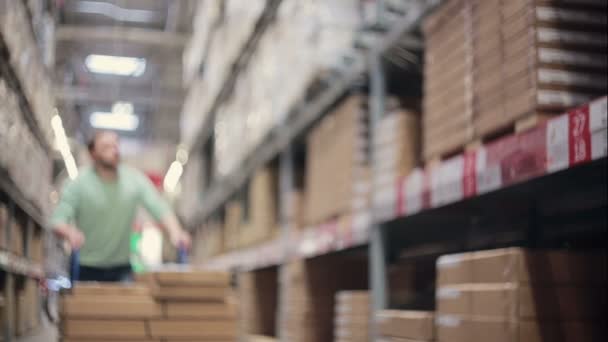 Un homme pousse un chariot entre des étagères avec des boîtes en carton dans un entrepôt de stockage — Video