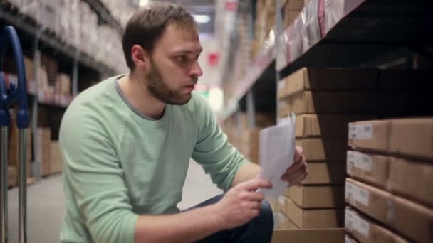 Un hombre está buscando los paquetes necesarios, sacándolos de la estantería con cajas de cartón en un almacén — Vídeo de stock