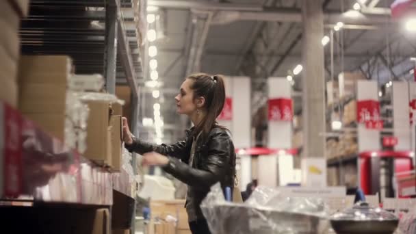 Mujer joven está de pie cerca de estantes con mercancías, en busca de la caja necesaria, sosteniendo en sus manos en un almacén — Vídeos de Stock