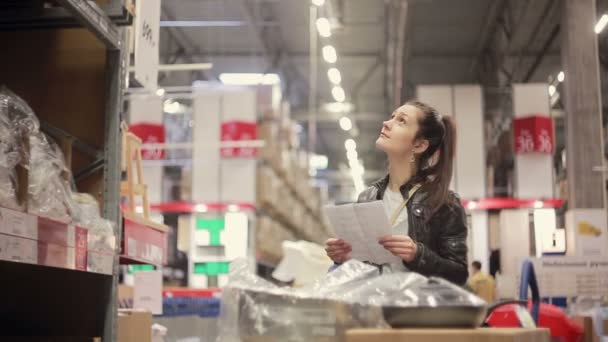 Een jonge vrouw is het controleren van haar lijst, op zoek naar iets op de planken, duwen haar trolley in een magazijn — Stockvideo