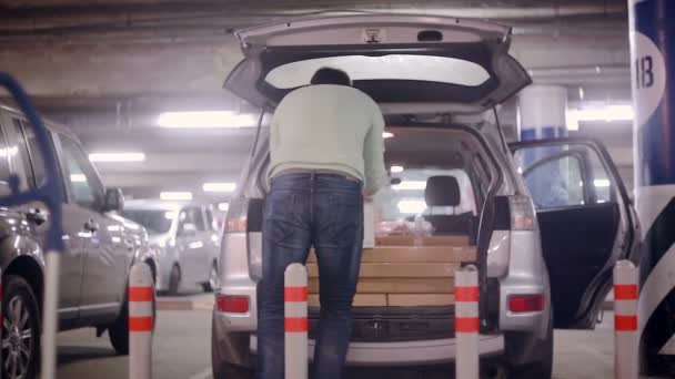 Un hombre está poniendo la compra en el maletero de un coche después de ir de compras de pie en el estacionamiento subterráneo cerca del centro comercial — Vídeos de Stock