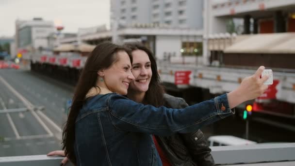 Две девушки лучшие друзья делают селфи, стоят на городском мосту, разговаривают, улыбаются, смеются. 4K — стоковое видео
