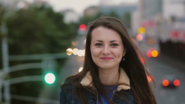 Vinden blåser långt hår vackra unga kvinnor. leende flicka står på bron på kvällen och ser på kameran. 4k — Stockvideo