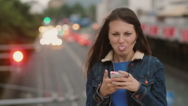 Весёлая, счастливая, улыбающаяся красивая женщина пользуется смартфоном, стоящим на мосту, развлекается, позируя смотрит в камеру. 4K — стоковое видео