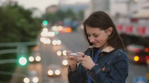 Happy, smiling pretty woman uses a smartphone standing on the bridge. Wind blows her hair. 4K — Stock Video