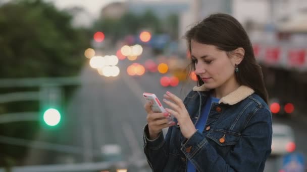 Mujer bonita feliz y sonriente utiliza un teléfono inteligente de pie en el puente. El viento sopla su cabello. 4K — Vídeo de stock