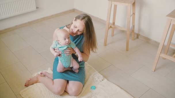 Young mother is holding her precious baby, smiling at him and laughing while sitting on the kitchen floor. Slow motion — Stockvideo