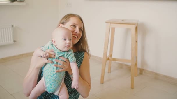 Joven madre está levantando a su precioso bebé, hablando con él con una sonrisa mientras está sentado en el suelo de la cocina cámara lenta — Vídeos de Stock